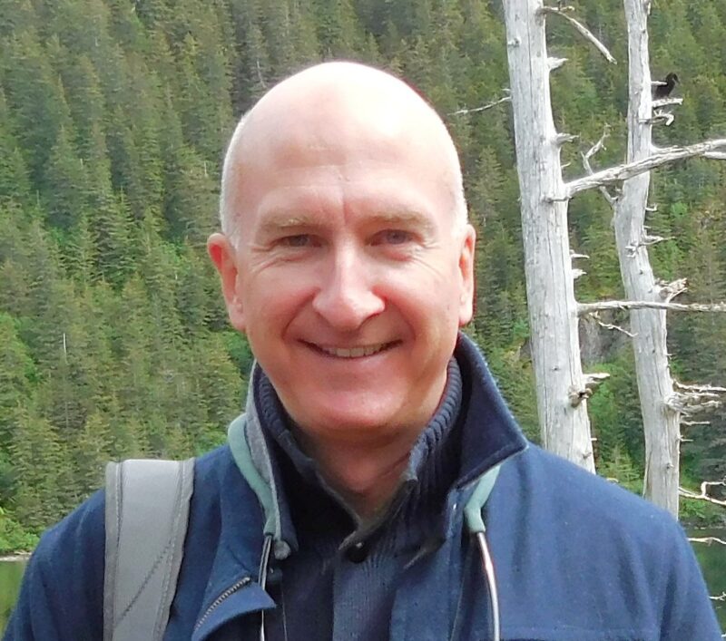 Casual headshot of Bill Connors in blue jacket with forest and trees in the background